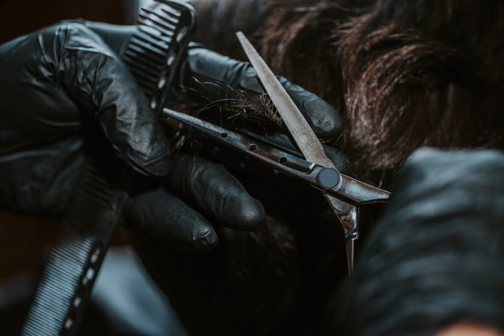 Mãos de luva preta segurando pente e tesoura, cortando uma mecha de cabelo escuro no topo da cabeça.