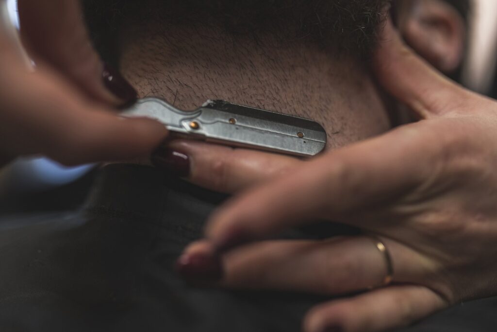 Mãos acertando com navalha o pelo curto da nuca de um cliente, com a gola da capa escura logo abaixo.