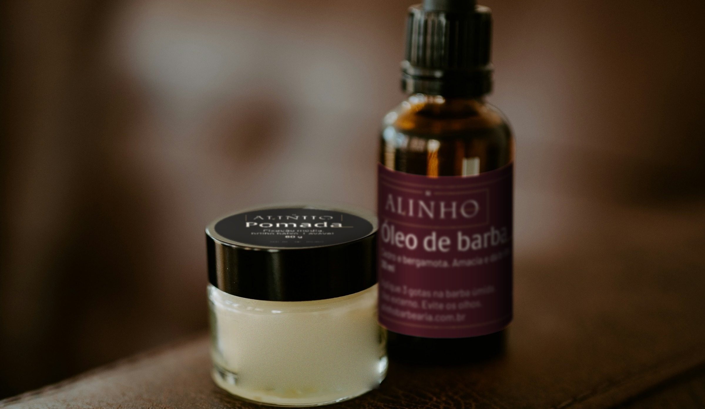 Frasco âmbar de óleo de barba Alinho, rótulo bordô, ao lado do pote de pomada de tampa preta, no braço de couro de um sofá.